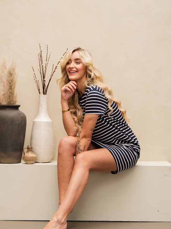 Vrouw in een marineblauw en wit gestreept T-shirtjurkje, zittend, met blonde krullen en tatoeages.