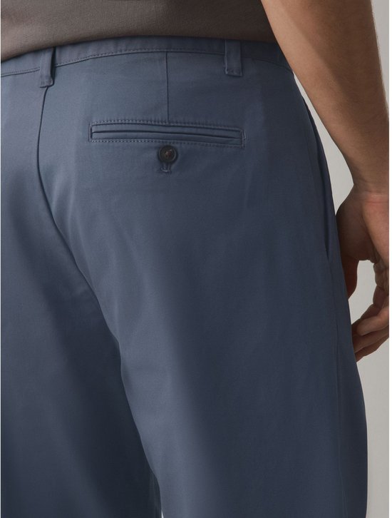 Close-up van de achterkant van een blauwe chino, met een detail van een klepzak met knoopsluiting.