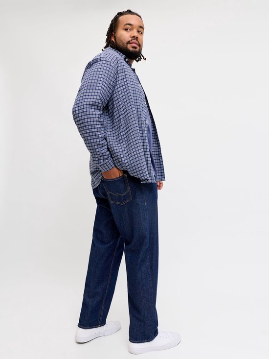 Man met dreadlocks draagt een blauw geruite blouse en donkere jeans, kijkend over zijn schouder.