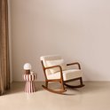 Schommelstoel met walnootkleurig houten frame, crèmekleurige bouclé bekleding en een bijpassend kussen. Naast de stoel staat een gestreepte bijzettafel met lamp.