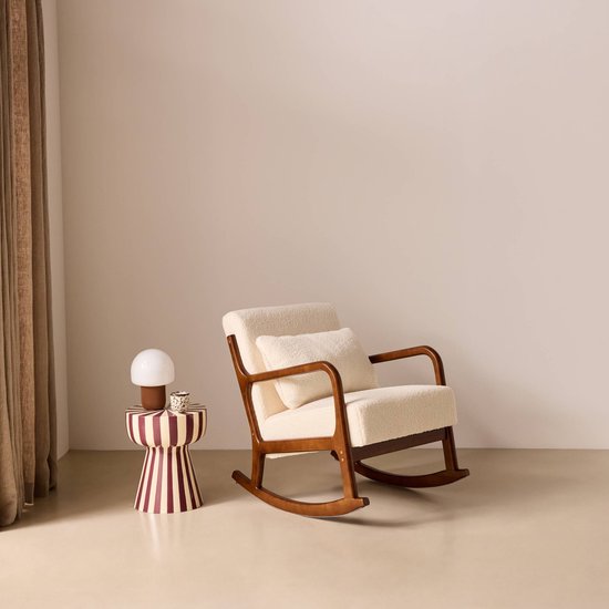Schommelstoel met walnootkleurig houten frame, crèmekleurige bouclé bekleding en een bijpassend kussen. Naast de stoel staat een gestreepte bijzettafel met lamp.