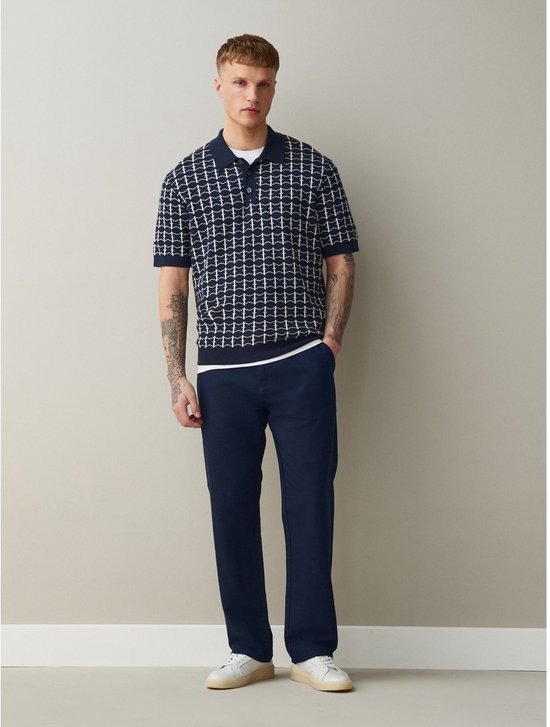 Man in donkerblauwe chino en polo met grafisch patroon, witte sneakers.