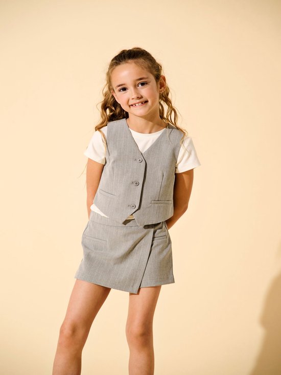 Meisje met lang golvend blond haar in een grijs gestreepte gilet, wit T-shirt en een bijpassende rok.