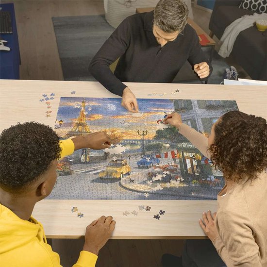 Drie personen zitten aan tafel en leggen een 2000-delige Ravensburger puzzel van Parijs bij zonsondergang.