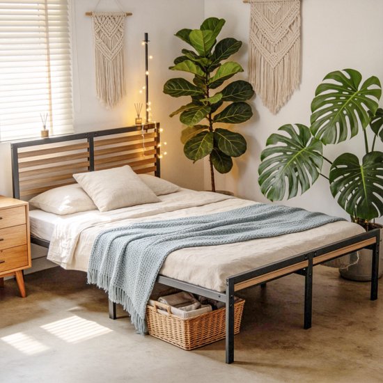Metalen bedframe met houten hoofdbord, gedekt met linnen beddengoed en een blauwgebreide sprei, rieten mand onderaan.