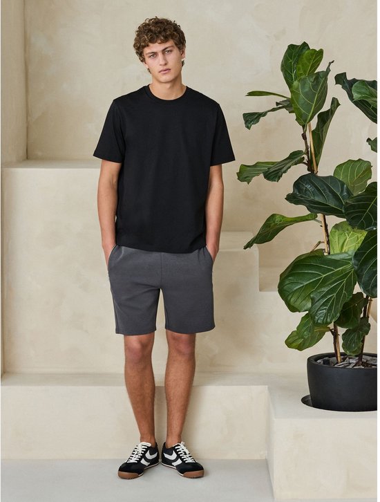 Model draagt een zwart T-shirt en grijze jersey short met ritszakken.