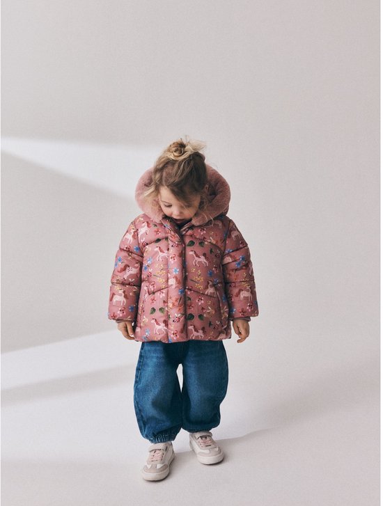 Kind in volledig roze gewatteerd jack met eenhoornprint, spijkerbroek en beige sneakers.