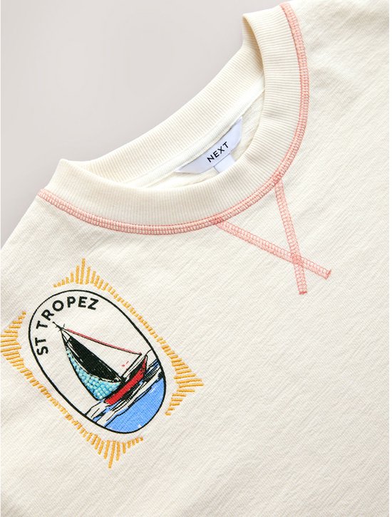 Crèmegekleurd T-shirt met ronde hals, roze stiksels en een gestikt embleem van een zeilboot met 'St Tropez'.