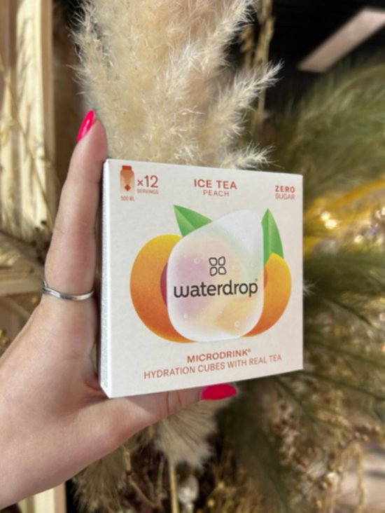 Hand met rode nagellak houdt een waterdrop Ice Tea Peach Microdrink doosje vast. Zicht op 12 servings.