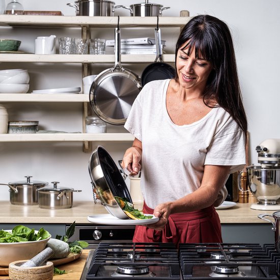 Vrouw met witte T-shirt en rode broek schept eten uit zilverkleurige pan op bord.