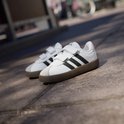 Twee witte sneakers met zwarte strepen, klittenbandsluiting en bruine zool op stenen ondergrond.
