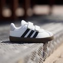 Witte sneaker met zwarte strepen, klittenbandsluiting en bruine zool.