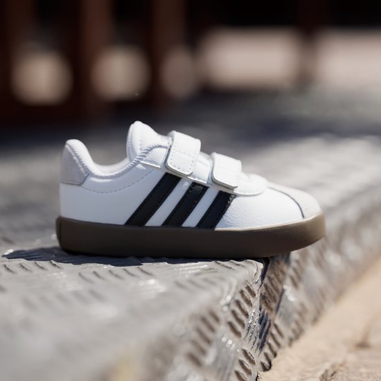 Witte sneaker met zwarte strepen, klittenbandsluiting en bruine zool.