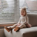 Een lachend blond meisje zit op een bankje bij een raam, met bruine Shoesme laarsjes aan.