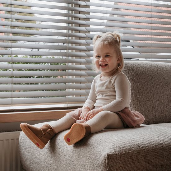 Een lachend blond meisje zit op een bankje bij een raam, met bruine Shoesme laarsjes aan.
