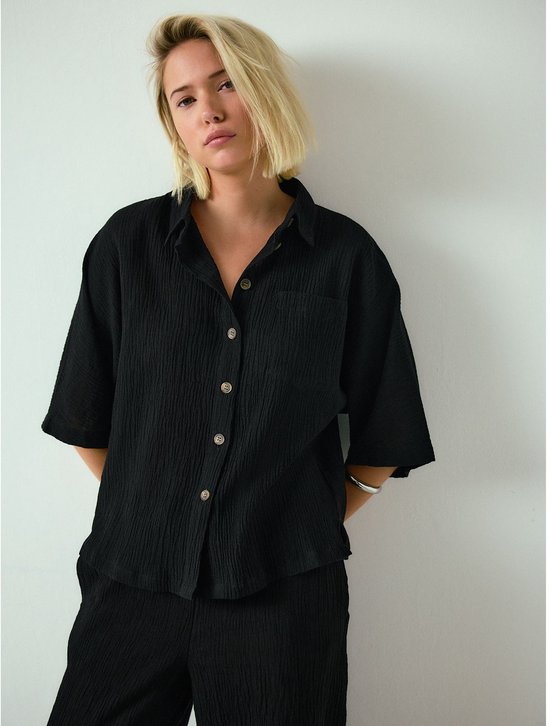 Model in zwart crêpe overhemd met korte mouwen en een wijde broek.