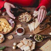 Een kersttafel met kaas, salami, olijven, crackers, jam, noten en een glas rode wijn.