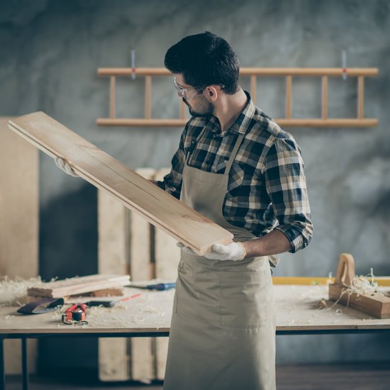 Een timmerman met veiligheidsbril en schort inspecteert houten planken in een werkplaats.