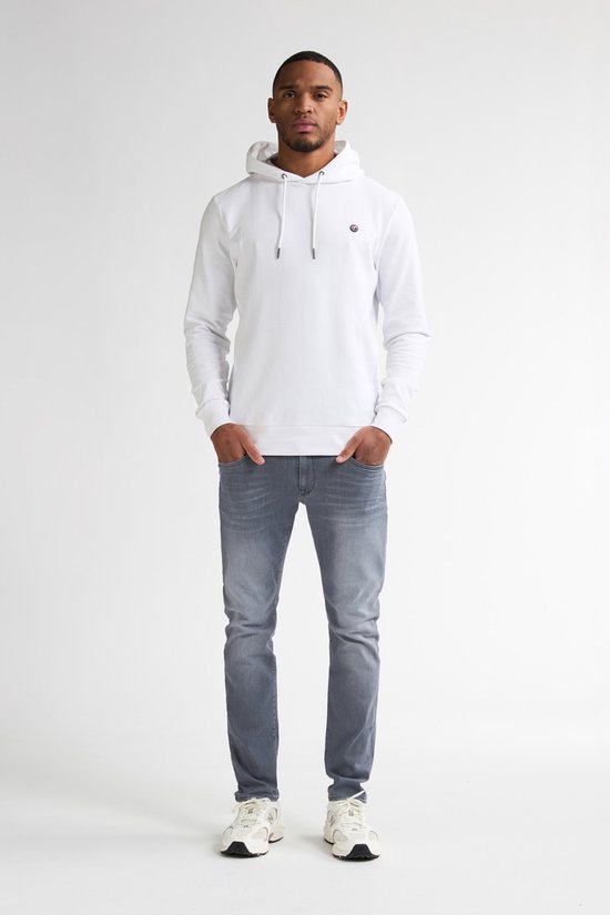 Man van voren gezien, draagt een witte hoodie en grijze slim fit jeans. Handen in de zakken.