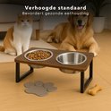 Dubbele hondenvoerbak op houten standaard, met twee RVS kommen. Een witte en een bruine hond zitten achter de voerbak. "Verhoogde standaard bevordert gezonde eethouding."