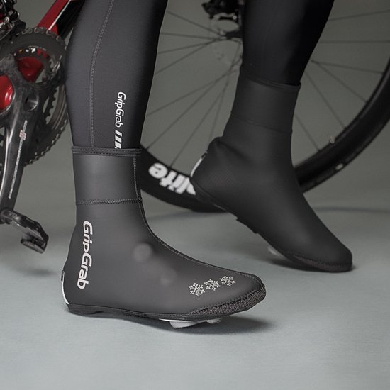 Persoon met zwarte fietsschoenen en "GripGrab" overschoudens, staand naast een racefiets.