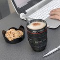 Zwarte cameralens beker met cappuccino en kaneel, naast koekjes en een laptop. Een hand typt op het toetsenbord.