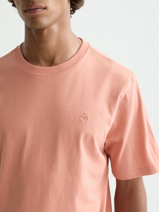 Model draagt een perzikkleurig T-shirt met korte mouwen en een geborduurd logo op de borst.