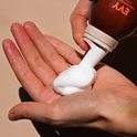 Hand met witte zelfbruinende mousse die uit een bruine EVY-flacon wordt gedrukt.