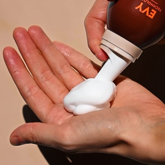 Hand met witte zelfbruinende mousse die uit een bruine EVY-flacon wordt gedrukt.