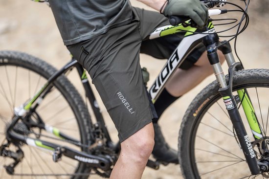Man op een mountainbike, close-up van de donkergroene korte fietsbroek met het logo "Rogelli" op het
been.