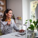 Een vrouw met een grijze trui drinkt koffie tijdens het werken op een computer.