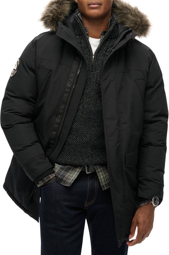 Man in zwarte Superdry parka met bontkraag, staand.