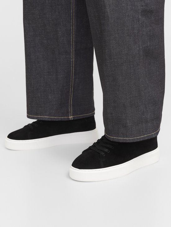 Persoon die zwarte suède sneakers met witte zolen draagt met een spijkerbroek.