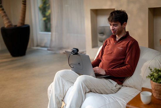 Man in fleece trui zit op een bank met een laptop, met webcam.