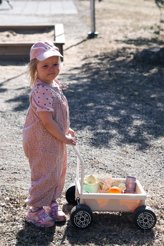 Een meisje met een roze pet en tuinbroek kijkt naar de camera terwijl ze een zandkleurige Plasto kinderwagen duwt.