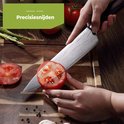Een hand snijdt een tomaat met een Hi Nature koksmes met damastpatroon en zwart handvat. Aspersies en tomaten liggen op de snijplank.