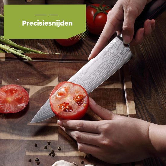 Een hand snijdt een tomaat met een Hi Nature koksmes met damastpatroon en zwart handvat. Aspersies en tomaten liggen op de snijplank.