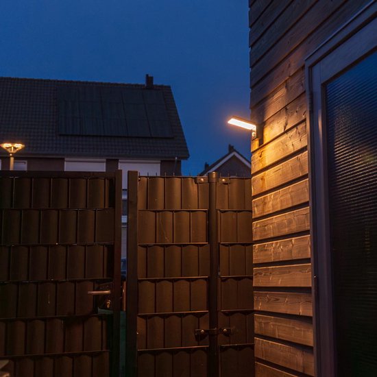 Zwarte wandlamp met sensor verlicht houten schutting naast een donkere poort in de avond.