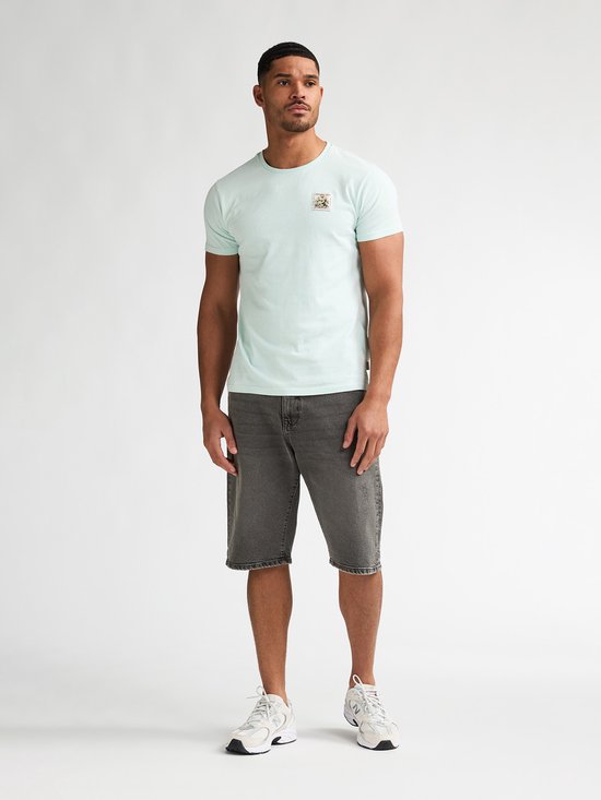 Man in lichtgroen T-shirt en grijze spijkerbroekshorts, van voren zichtbaar.