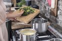 Een persoon legt een houten snijplank met broccoli boven twee RVS pannen op een gasfornuis.