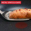Een stuk zalmfilet met peperkorrels, op een metalen spatel, boven een zwarte koekenpan.