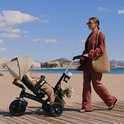 Vrouw in roze pak duwt een beige driewieler met kind op het strand.