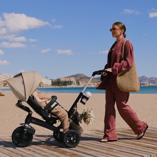 Vrouw in roze pak duwt een beige driewieler met kind op het strand.