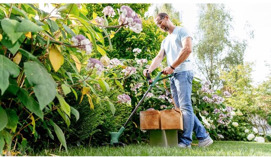 Man met blauw shirt gebruikt de Bosch grastrimmer in een tuin met bloeiende struiken.