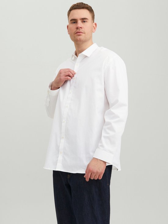 Man draagt een wit Jack & Jones overhemd en donkere jeans, met zijn hand op het overhemd.