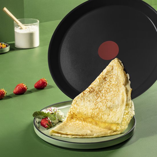 Zwarte koekenpan met een opgevouwen crêpe, versierd met aardbeien, munt en slagroom op een groen bord.
