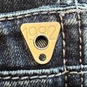 Detail van een gouden driehoekige knoop met '1982' erop, op een blauwe jeans.