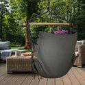 Donkergrijze hangstoel met spreidstok en franjes op een houten terras bij een tuin.