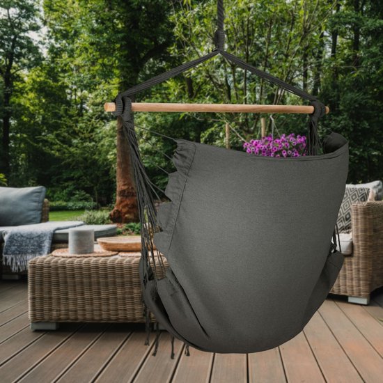Donkergrijze hangstoel met spreidstok en franjes op een houten terras bij een tuin.