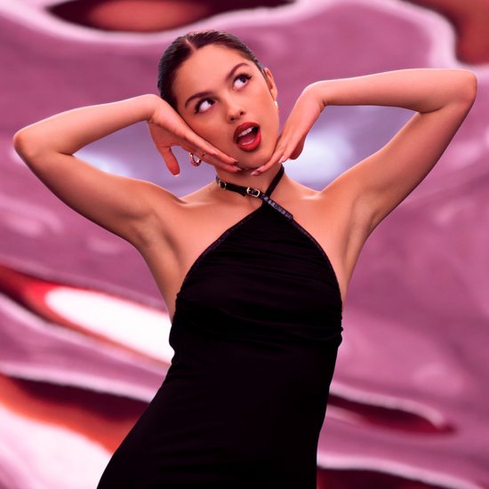 Model met handen op gezicht, draagt zwarte halterjurk en zwarte choker, roze achtergrond.